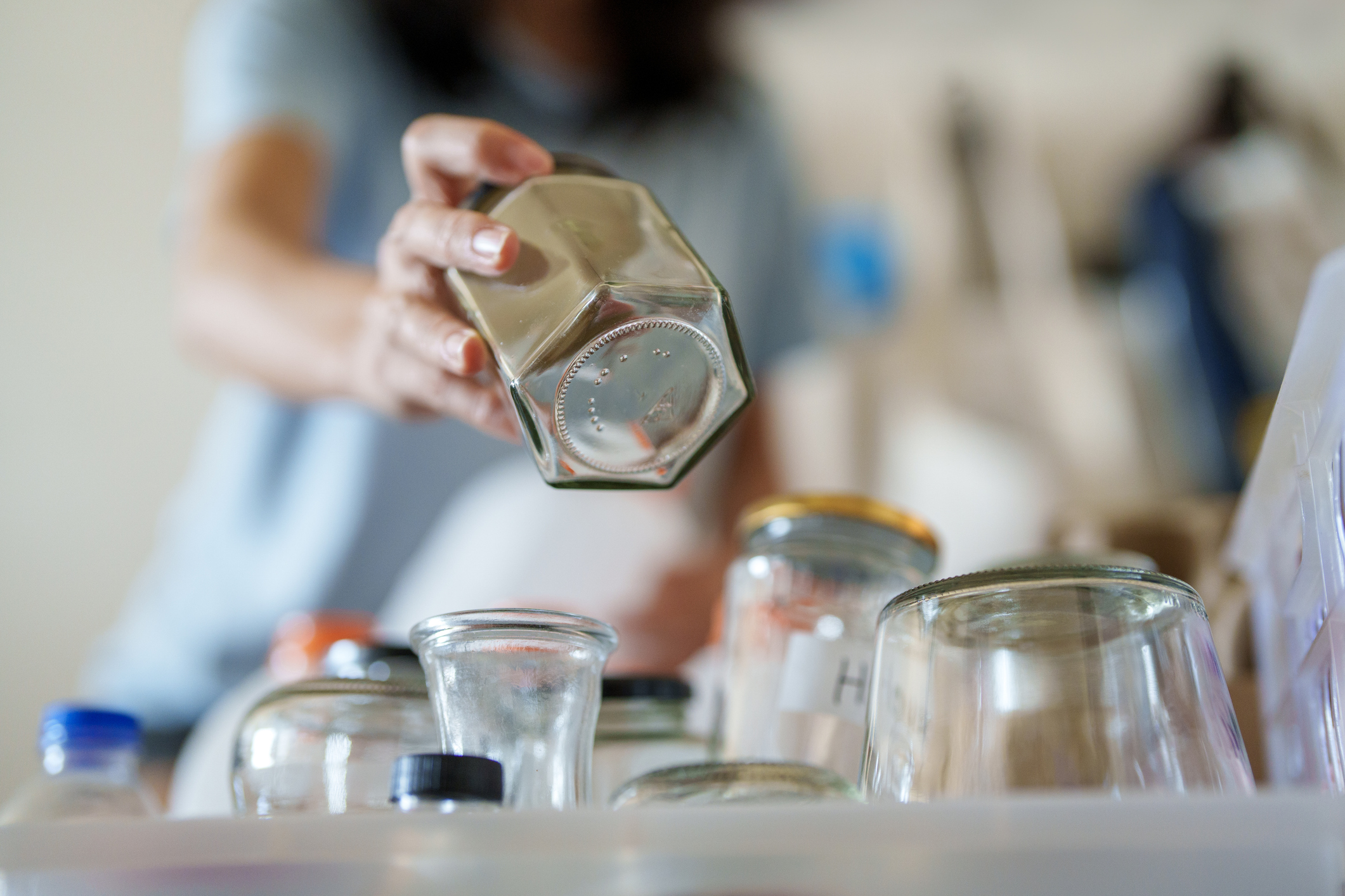 A woman placing an empty glass jar onto a large, cluttered stack of similar glass jars in a kitchen cabinet.