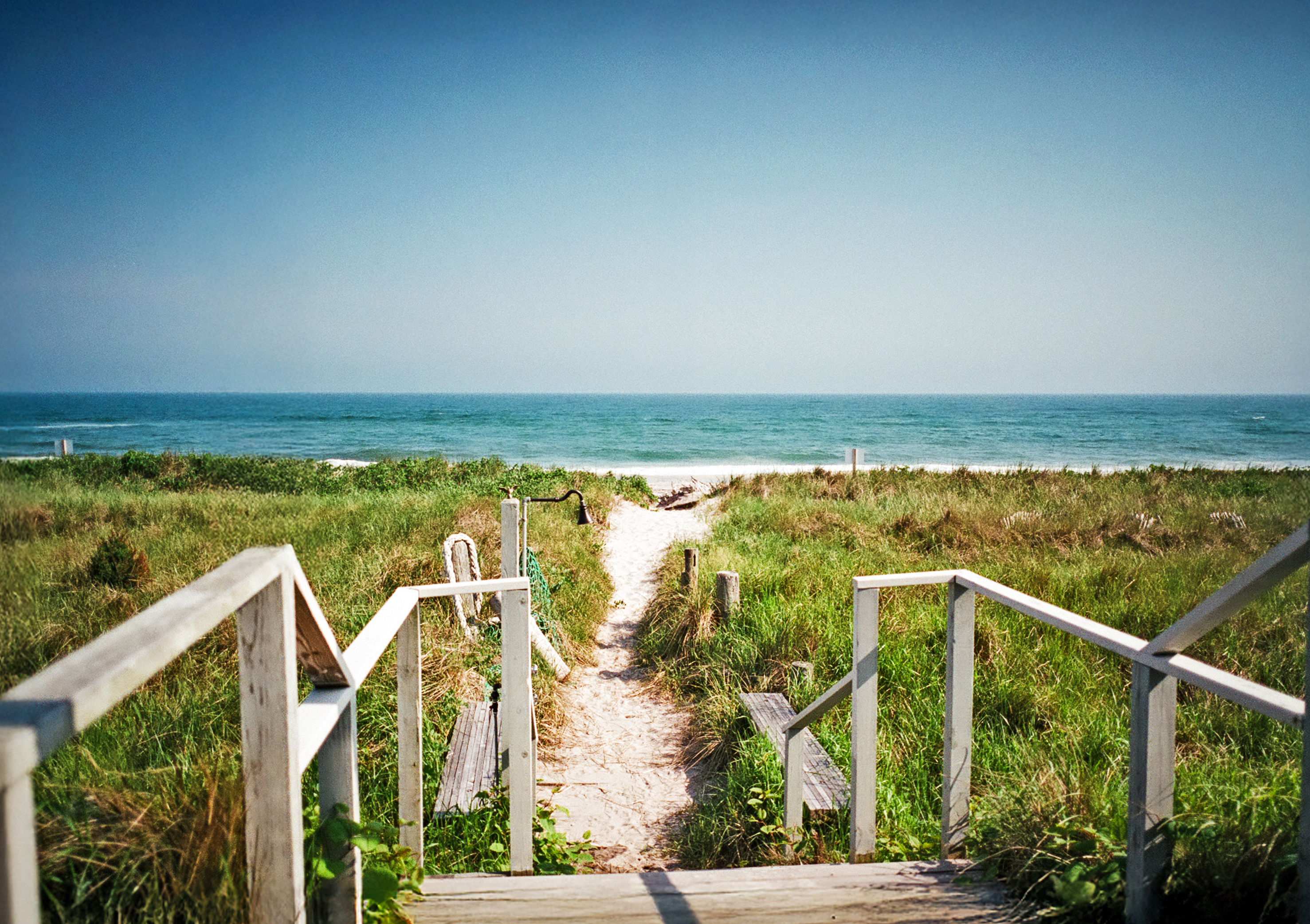A peaceful view of a Hamptons sandy beach seen from a wooden porch balcony.