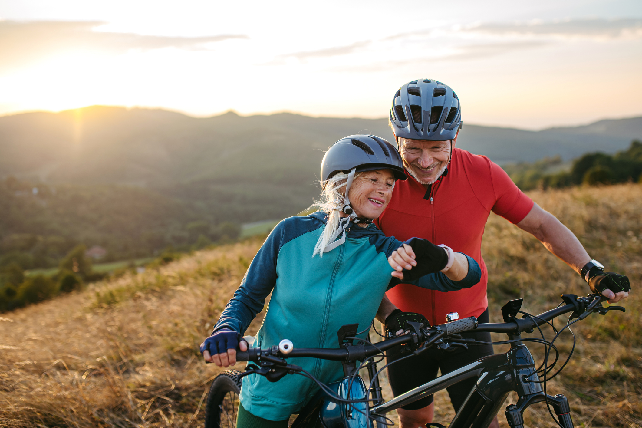 An active older couple smiling while mountain biking outdoors in a scenic area.