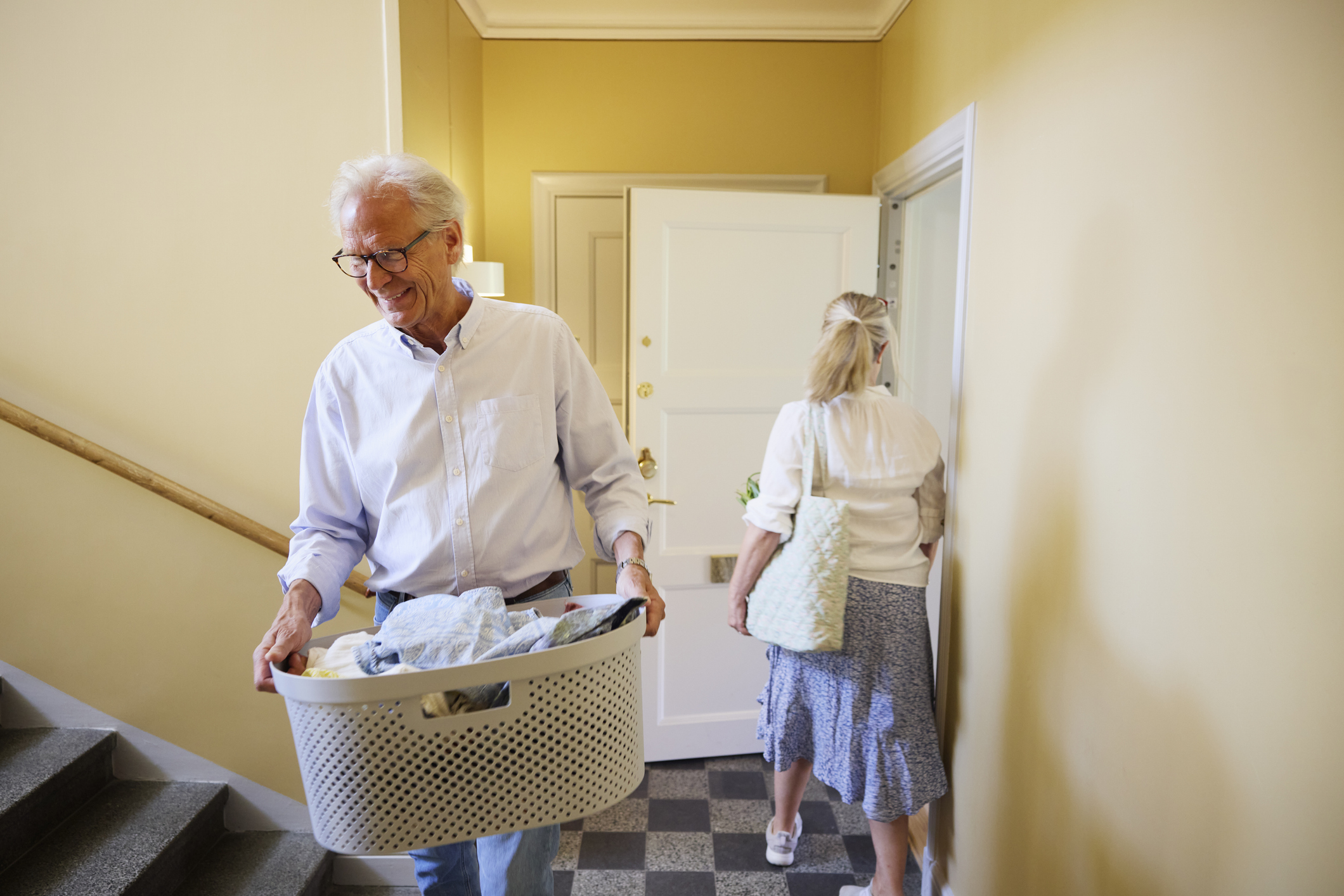 A mature couple in a home laundry room carrying a basket of clothes.