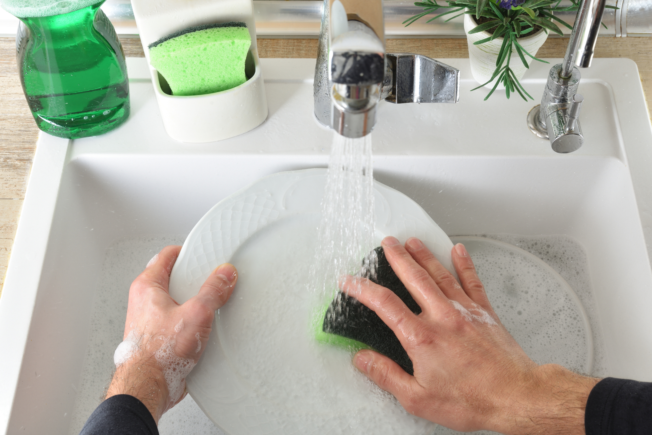 Close-up of a person's hands washing a white ceramic plate under a stream of running water in a kitchen sink.
