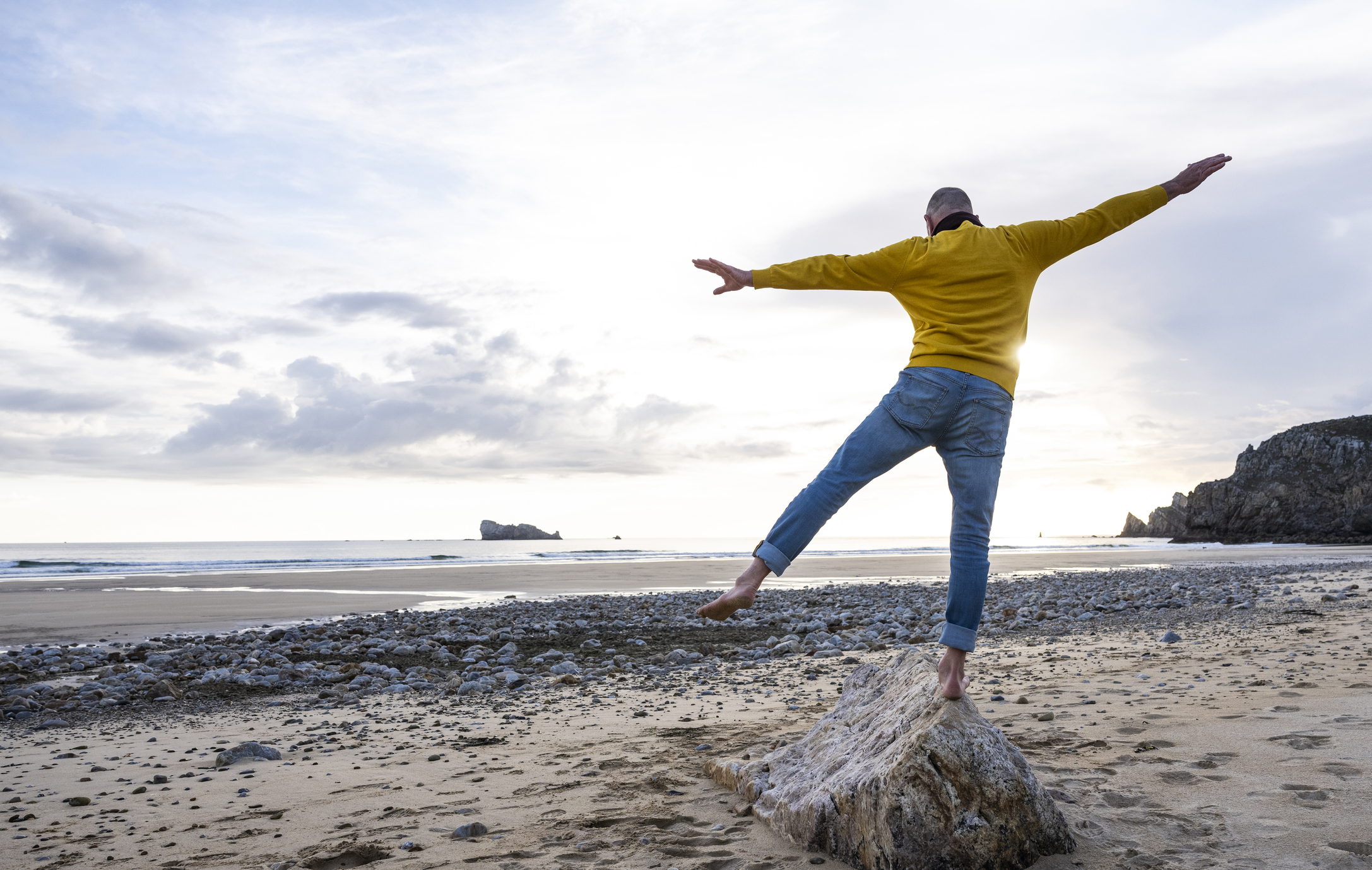 An active older man in a yellow shirt balancing on one leg on a beach rock.